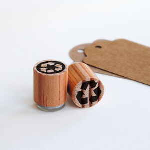 K&ouml;nnte beinhalten: Zwei Holzstempel mit schwarzen Recycling-Symbolen. Die Stempel befinden sich auf einer wei&szlig;en Oberfl&auml;che.