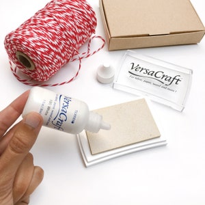 May include: A white ink pad with a clear plastic case that says "VersaCraft for fabric, paper, wood and more!"  A white bottle of VersaCraft ink labeled "180 White 1 fl oz (30ml)" is being held in a hand.