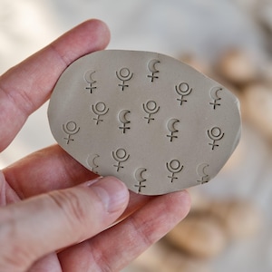 Könnte beinhalten: Ein hellbeige, oval geformter Tonstempel mit einem Muster aus Himmelszeichen, darunter Halbmonde und Planetensymbole. Der Stempel wird in einer Hand gehalten, wodurch die detaillierten Eindrücke zur Geltung kommen. Der Ton hat eine glatte, matte Oberfläche.