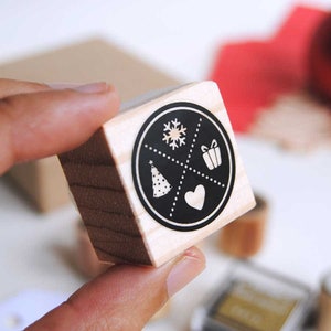 Puede incluir: Un sello de madera con un diseño circular que presenta un copo de nieve, un regalo, un corazón y un árbol de Navidad. El sello se sostiene en una mano, con un fondo borroso de otros suministros de artesanía.