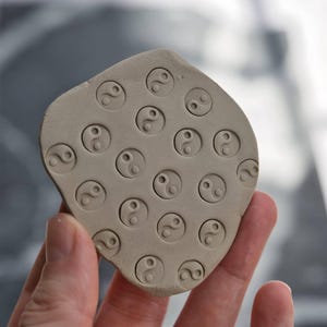May include: A light beige clay stamp with multiple Yin Yang symbols pressed into the surface. The stamp is held in a hand, and the background is blurred.
