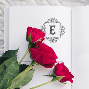 The Personalized Ex Libris Moody Blooms stamp is printed on an open book with three red roses resting gently on the pages. The rich floral lines and monogram stand out in black ink, evoking gothic romance, fantasy literature, and poetic charm. A beautiful way to mark your own romantic fantasy book collection or create a memorable gift for a fellow literary dreamer.