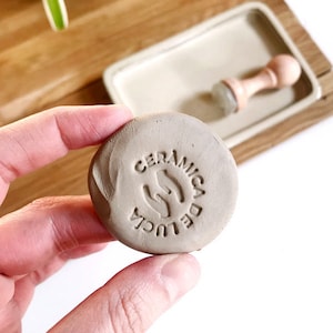 May include: A close-up of a brown clay stamp with the text "CERAMICA DE LUCIA" and a hand symbol in the center.