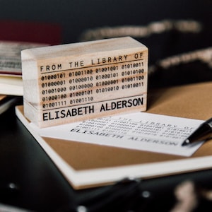 Könnte beinhalten: Ein Holz-Bibliotheksstempel mit dem Text "FROM THE LIBRARY OF" und dem Namen "ELISABETH ALDERSON". Der Stempel enthält Binärcode. Der Stempel liegt auf einer weißen Karte und einem braunen Notizbuch.