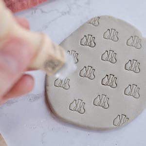 Può includere: Una lastra di argilla grigio chiaro con un motivo ripetuto di un design di vaso stilizzato. Una mano preme un timbro sull'argilla, creando le impressioni. Lo sfondo è una superficie di marmo bianco.
