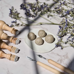 May include: Four small clay balls with different facial expressions on a white plate.  There are five wooden stamps with the text "bier gut" on them and two wooden tools on a white marble surface.