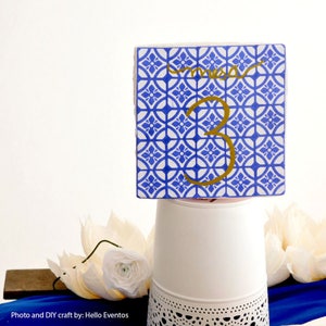 May include: Blue and white patterned tile with a gold number 3 and the word "mesa" written in gold cursive. The tile is sitting on a white stand with a lace pattern.