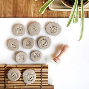 May include: A collection of nine round clay stamps with various designs, including a heart, a bird, initials, and the words "Pottery By" and "Martha Geller". A wooden stamp with the word "Goodnight" is also included.