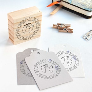 May include: A wooden stamp with a circular design featuring a calendar with the text "LAID ON" and a chicken illustration. The stamp is used to mark dates on paper tags.