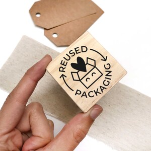 May include: A wooden stamp with a black ink design of a box with a heart inside and the text "REUSED PACKAGING" in a circle around it.