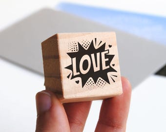 Könnte beinhalten: Ein quadratischer Holzstempel mit einem schwarzen Grafikdesign in Form einer Comic-Sprechblase, in deren Mitte das Wort LOVE gedruckt ist, umgeben von kleinen Punkten und Herzformen auf der hellbraunen Holzoberfläche.