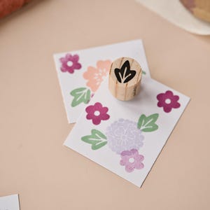 Peut inclure: Un tampon en bois avec un motif de feuille est posé sur deux cartes décorées de motifs floraux roses, violets et verts. Le tampon est cylindrique et a une finition en bois naturel. Les cartes présentent des fleurs et des feuilles estampillées.