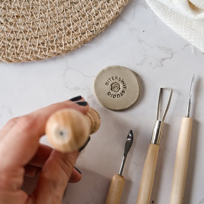 May include: A close-up of a clay stamp with the text "Bittersweet Studio" and a rose design. The stamp is on a white surface with a wooden handle and three metal tools.