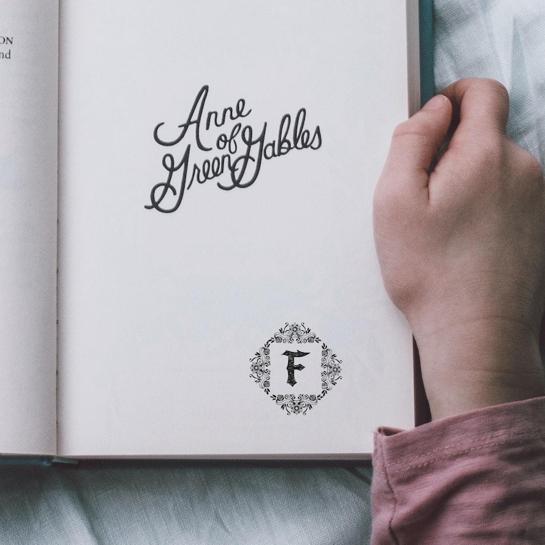 Close-up of a hand holding an open copy of Anne of Green Gables, showing the floral monogram library stamp printed on the bottom right corner of the page. The detailed bloom design blends beautifully with the nostalgic charm of the book. Ideal for book lovers who want to mark their collection with elegance and old soul vibes. Great for dark romanticism fans and personalized bookplate lovers.