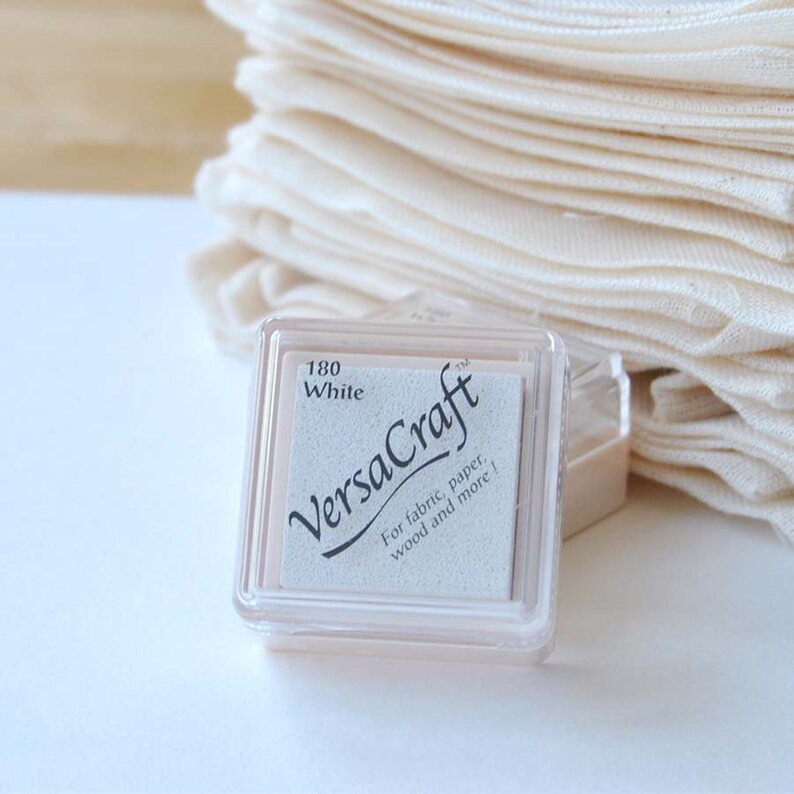 May include: A white ink pad in a clear plastic case. The ink pad is labeled "VersaCraft" and "180 White". The label also says "For fabric, paper, wood and more!"