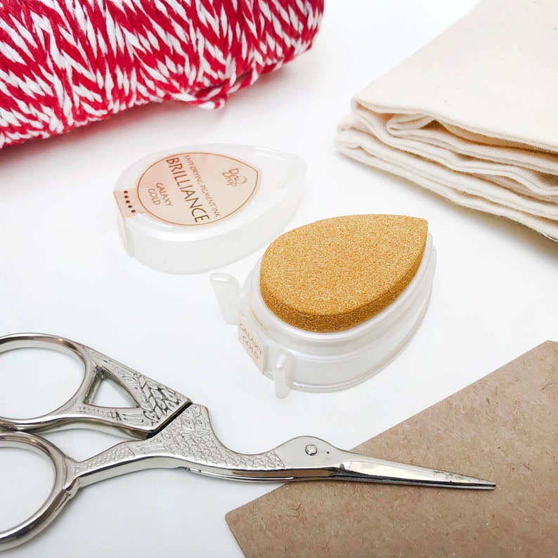 May include: A close-up of a gold ink pad with a sponge applicator. The ink pad is labeled "BRILLIANCE" and "GALAXY GOLD" and is in a white plastic container. The ink pad is next to a pair of silver scissors with a bird-shaped handle.