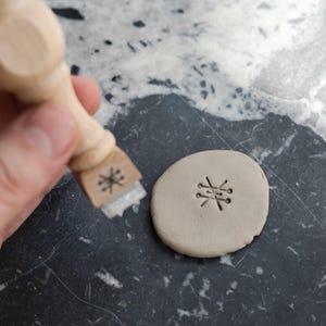 May include: A light-colored wooden stamp with a geometric starburst design on the base, positioned next to a circular piece of light gray clay that features the same starburst pattern impressed into its surface.