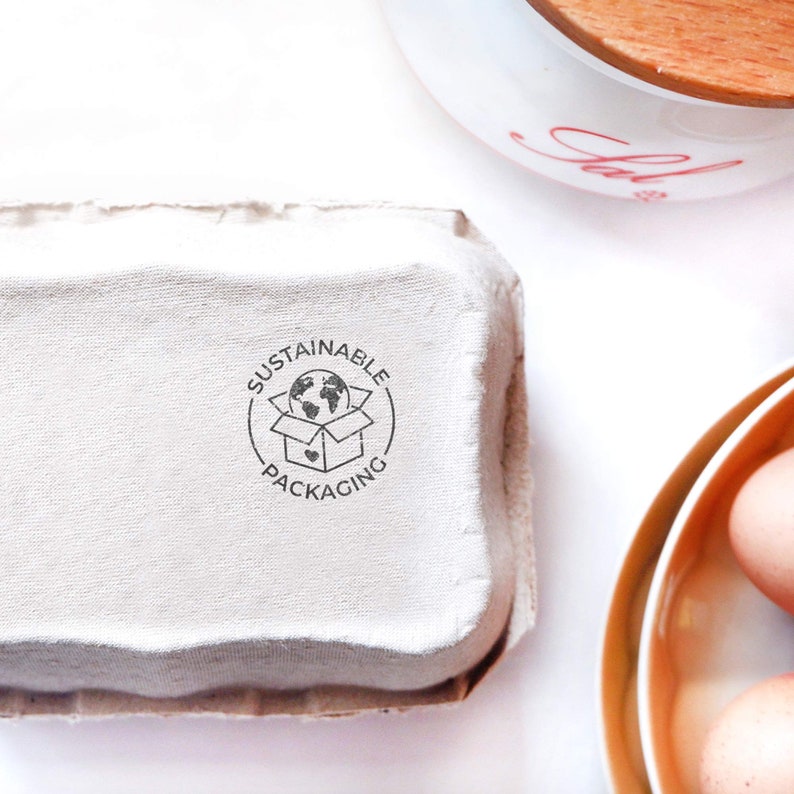 May include: A white cardboard egg carton with a black and white stamp that reads "Sustainable Packaging" with a globe inside a box.