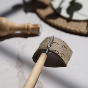 May include: A close-up of a clay piece with a leaf pattern being smoothed with a wooden tool. The clay is a light brown color and the tool is a light brown color with a silver metal tip.