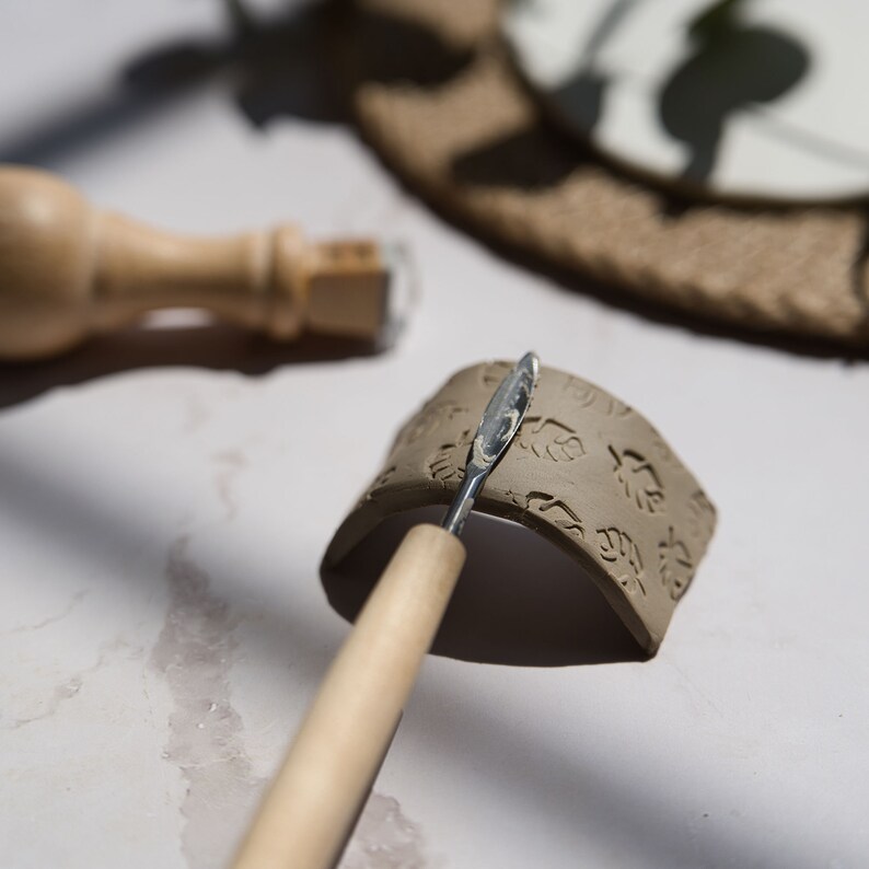 May include: A close-up of a clay piece with a leaf pattern being smoothed with a wooden tool. The clay is a light brown color and the tool is a light brown color with a silver metal tip.