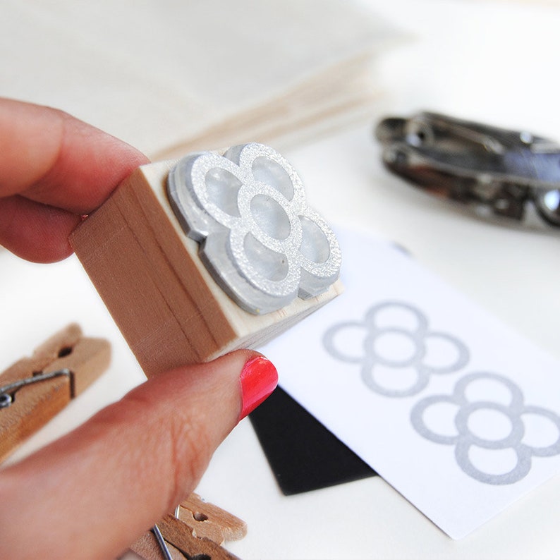 May include: A wooden stamp with a silver metal design of a flower with six petals. The flower has a textured surface. The stamp is being held by a hand with red nail polish.