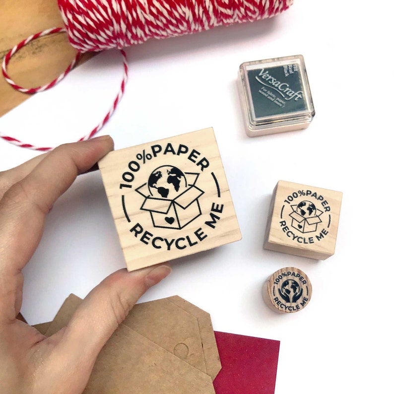 A hand holds the M-size 100% Paper Recycle stamp, displaying the larger surface area for clear and bold impressions. The wooden mount offers a sturdy grip for effortless stamping on paper bags, mailers, and shipping boxes. This size is perfect for businesses needing a visible, eco-conscious label on their sustainable packaging materials.