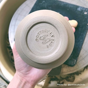 May include: A close-up of a light brown ceramic bowl with a hand-stamped logo that reads "Pottery by Jade" on the bottom.