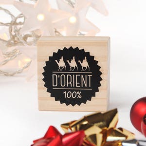 May include: A wooden stamp with a black and white design of three people riding camels. The stamp says "D'ORIENT 100%".