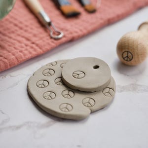 Peut inclure: Ornements artisanaux en argile avec le symbole de la paix. Les ornements sont gris clair et présentent le symbole de la paix. Un outil en bois avec le symbole de la paix est en arrière-plan, ainsi que d'autres outils d'artisanat et un tissu rose.
