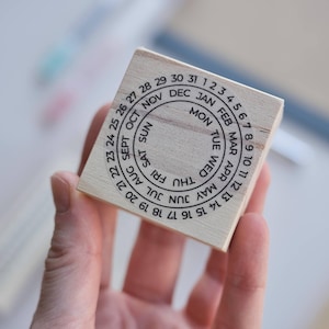 Pode incluir: Um carimbo de madeira quadrado com um design circular e texto impresso em preto. O anel externo exibe os números de 1 a 31, o anel central lista os meses de JAN a DEC e o círculo interno contém os dias da semana de MON a SUN.