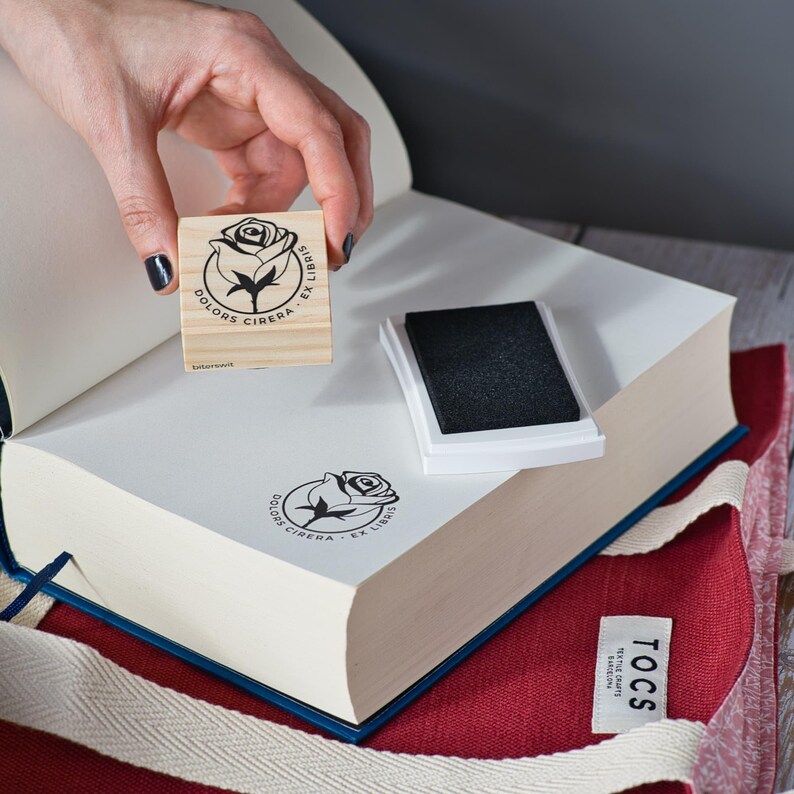 A hand is shown mid-stamp, pressing the rose-themed ex libris onto an open book page using black ink. The stamp, part of Biterswit’s floral collection for readers, is perfect for journaling, custom bookplates, or gifts for moms and teachers who adore books. Ideal for personal libraries, reader moms, and romantic floral design lovers who want to add handmade flair to their pages.