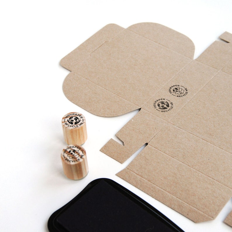 Close-up of mini rubber stamps printed on a cardboard box, demonstrating how the "100% Paper Recycle" stamp enhances eco-conscious packaging. The crisp imprint on recyclable material highlights the importance of reducing plastic waste in shipping. This small yet impactful tool is perfect for brands committed to sustainability and plastic-free packaging.