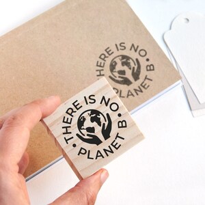 A medium-sized rubber stamp featuring hands holding the Earth is held in hand, showing its design stamped onto a notebook cover. This image highlights how the stamp can be used beyond packaging—on journals, planners, and DIY projects. The hands design symbolizes care for the planet, making it a great tool for small businesses, eco-conscious brands, and crafters looking to personalize their products while promoting a sustainability message.