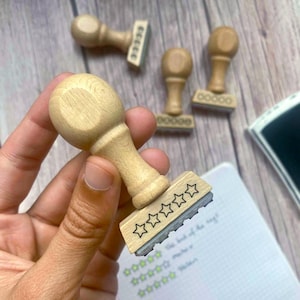May include: Wooden stamps with star designs. One stamp is held in a hand, with four others in the background. An ink pad is visible on the right. The stamps are for rating or review purposes.