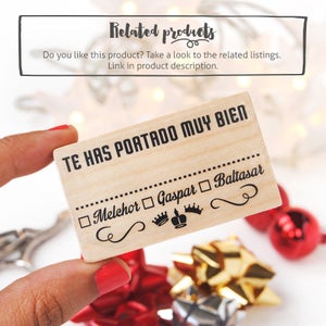 May include: Wooden stamp with the text "Te has portado muy bien" (You have behaved very well) and a checklist with the names of the three Wise Men: Melchior, Gaspar, and Baltasar. The stamp is held in a hand against a background of red and gold gift wrap.
