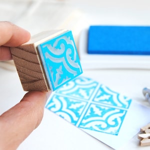 May include: A wooden stamp with a blue and white patterned design. The stamp is being held in a hand and is about to be used to create a print.