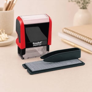 May include: A Trodat Printy 4911 typo self-inking stamp with a red and black plastic casing. The set includes a black plastic base containing a grid of small rubber characters and a pair of black tweezers for arranging the letters on the stamp.