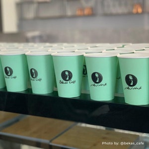 May include: A row of light green disposable cups with a black logo that says "B&eacute;kas Caf&eacute;" and a stylized image of a person's head. The cups are lined up on a black shelf.