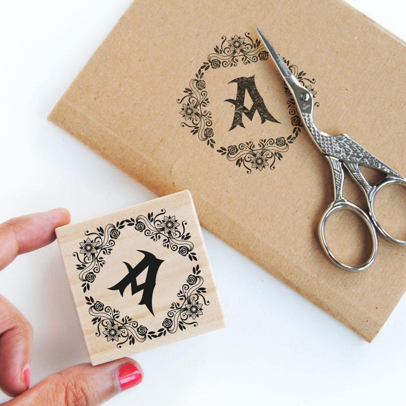 A hand holds the romantic floral monogram library stamp in a 5 cm pine wooden block while it’s stamped on a kraft notebook cover. Beside it, vintage swan-shaped scissors add to the dark romantic stationery vibe. The stamp's floral details and initial shine through in deep black ink, giving the notebook a personalized and gothic touch. Perfect for literary girl journaling, romantic bookish gifts, and dark academia-inspired book accessories.