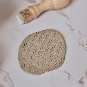 Könnte beinhalten: Ein Holzstempel mit floralem Design liegt neben einem Tonstück mit geprägtem Muster. Der Stempel hat einen Holzgriff und einen quadratischen Stempelkopf. Der Ton ist oval geformt und hat ein sich wiederholendes blütenartiges Muster.