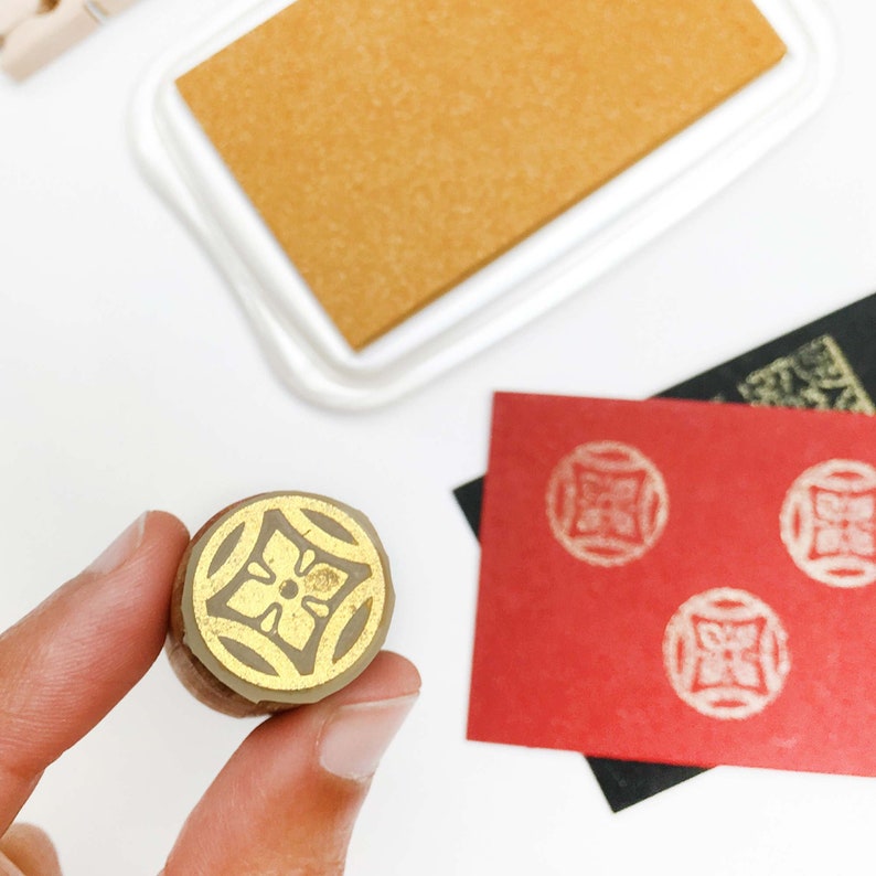 May include: A close-up of a wooden stamp with a gold floral design. The stamp is being held by a hand. The stamp is used to create a decorative design on paper.