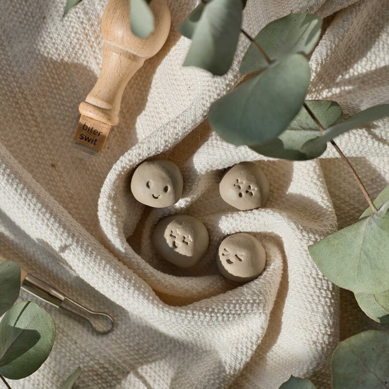 May include: Four small, beige, clay-like ornaments with different facial expressions. The ornaments are on a white fabric background with green leaves.