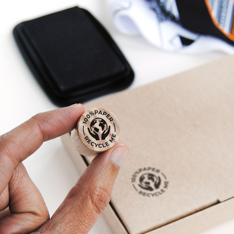 A hand holds the mini rubber stamp, showing its compact size and eco-friendly wooden block. The stamped design on the paper box reads "100% Paper Recycle," encouraging sustainable packaging. This stamp is ideal for branding paper envelopes, cardboard, and recyclable packaging materials, making it perfect for small businesses and zero-waste shops.