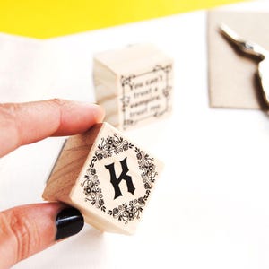 A hand displays the smaller version of the Regal Fantasy Bookplate on a plain white background. In the blurred background, another stamp reads don’t trust a vampire trust me, setting a mysterious Halloween tone. This moody floral monogram stamp is ideal for readers who love romance fantasy, dark and edgy stationery, or personalized ex libris stamps with a twist of fantasy lore.