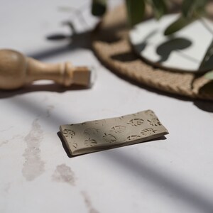 May include: A close-up of a rectangular piece of clay with a pattern of leaves pressed into the surface. The clay is a light brown color.