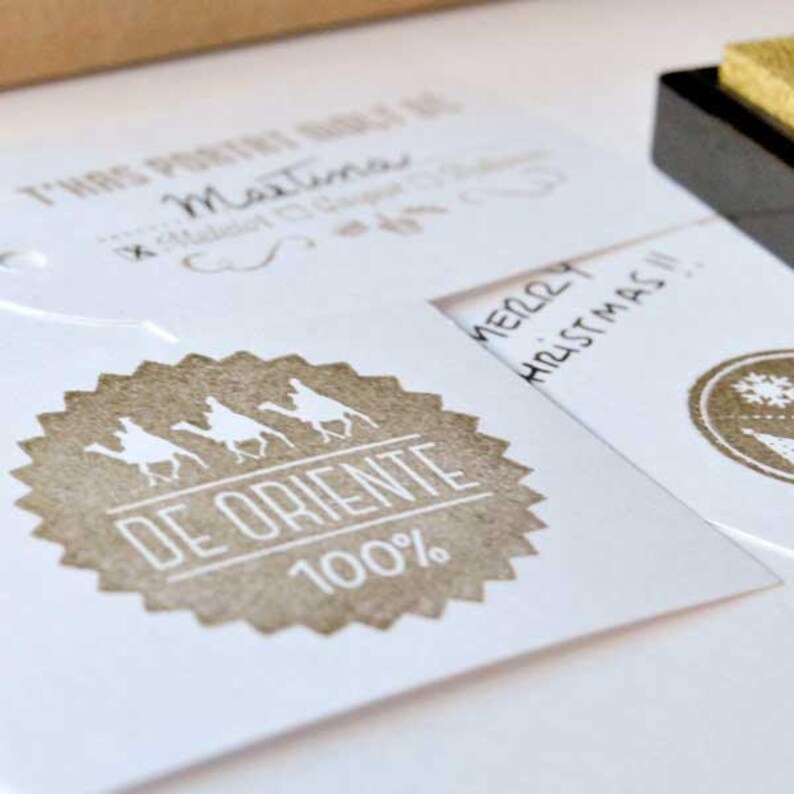 May include: A white card with a brown circle stamp featuring three figures riding camels with the text "DE ORIENTE 100%". The card also has the text "Merry Christmas!" in cursive.