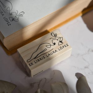 A vintage open book lies beside two stamps: the floral monogram stamp and a minimal line drawing ex libris with a woman silhouette. Both designs speak to different aesthetics—moody romanticism and minimalist elegance—making them perfect for book lovers with a taste for custom, meaningful stationery. A lovely duo for gift-giving or creating your own bookish ritual.