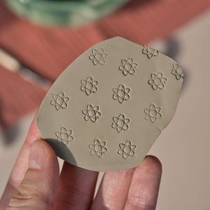 Puede incluir: Un pequeño sello de arcilla gris de forma irregular con un patrón repetitivo de diseños florales en forma de átomo. El sello se sostiene en una mano, con un fondo borroso de una superficie marrón rojiza.