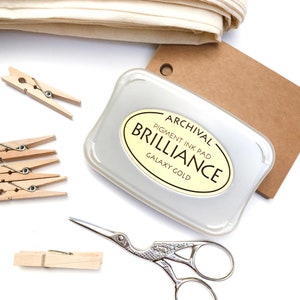 May include: A silver ink pad with a yellow label that reads "Archival Pigment Ink Pad Brilliance Galaxy Gold". The ink pad is on a brown paper surface.
