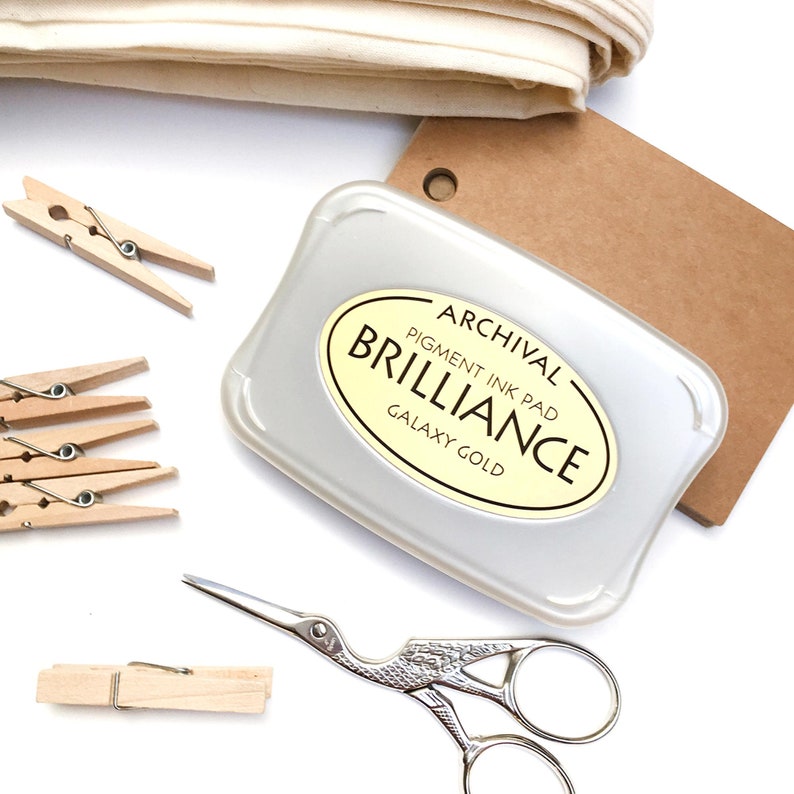 May include: A silver ink pad with a yellow label that reads "Archival Pigment Ink Pad Brilliance Galaxy Gold". The ink pad is on a brown paper surface.